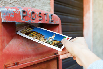 Male hand is putting a postcard in a red mailbox in Florence (Firenze), Italy. The postcard shows a...
