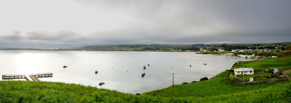 A wide, panoramic view of Petre Bay, Chatham Islands, New Zealand, on a dark, overcast day. Waitangi township on the right, and Waitangi Wharf on the left. 