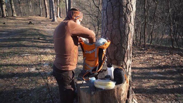Theme hiking and travel. A Caucasian tourist man unpacks an orange backpack, takes out his things and puts them on a stump in the forest. Equipment and things for camping