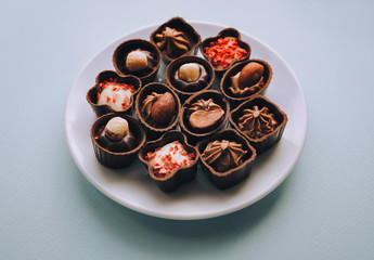 Beautiful chocolates of different shapes and fillings lie in a white plate against the blue background paper. Close up.
