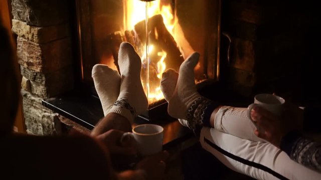 4K Couple seating near fireplace stretching their legs having warm beverages. Wearing Christmas socks.