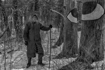 A man in a death suit with a scythe: a black cloak with a hood, a mask of a skull and a scythe in his hand, somewhere in forest. Maybe halloween costume. Black and white image