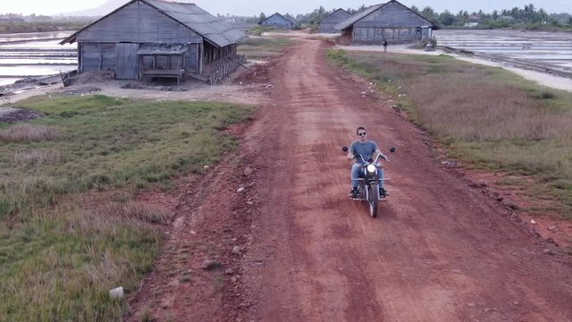 Aerial drone view tracking in front of a motorcycle rider as he rides slowly offroad in Asia.  Old wooden sheds and shacks can be seen.