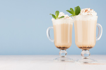 Two coffee drinks with whipped cream, green mint, cocoa powder for breakfast in glass with handle in  pastel blue interior on white wood table, copy space.