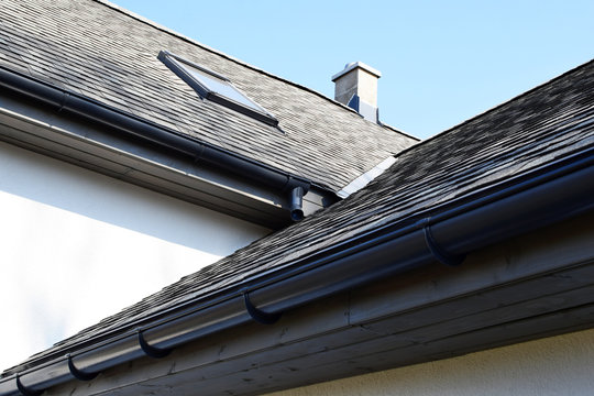 Grey asphalt shingles roof construction with ridge ventilation on rooftop, wooden eaves with plastic rain gutter, window and chimney, copy space on white wall or blue sky.
