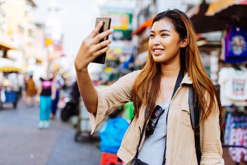 Young Asian female tourist woman taking selfie photos with smartphone while travelling in Bangkok,...