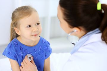 Fototapeta premium Doctor examining a little girl by stethoscope. Happy smiling child patient at usual medical inspection. Medicine and healthcare concepts