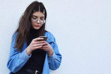 Pretty teen girl using smartphone for chatting and texting outdoors on white plaster wall background, brunette wearing blue jeans shirt and stylish round glasses, copy space.