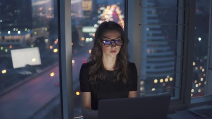 young business woman working late in the office. portrait of a girl with glasses on the background of the night city - Powered by Adobe