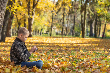 Profile portrait of white kid of 10 years sitting in golden autumn park on ground and using his smart phone. Horizontal color photography.