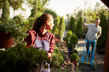 Fototapeta na wymiar Girl gardener takes care of the plants while the guy rolls the cart on the garden path on a sunny day. Working in the garden