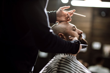 Fashion barber dressed in a black clothes tidies up men's beard  and scissors it in the barbershop