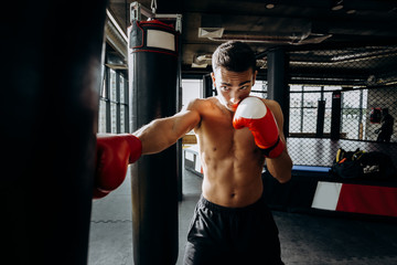 Sportsman in red boxing gloves with a naked torso hits punching bag in the gym on the background of...