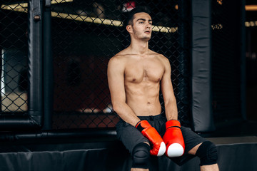 Sportsman with a naked torso and the red boxing gloves  on his hands  sits on the border of boxing...