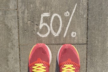 Text 50% written on gray pavement with woman legs in sneakers, view from above