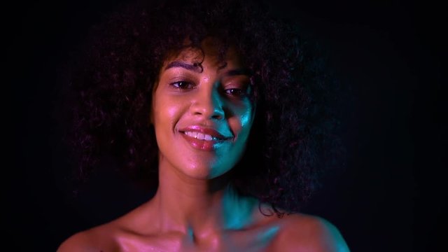Portrait of young seductive african american girl in pink neon light at black background. Tempting woman with perfect makeup looking at camera and smiling. Glamour, fashion concept.