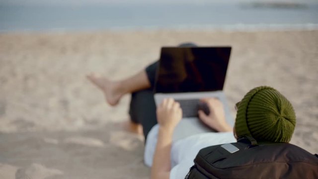 Close up behind shoulder shot of young professional freelancer or tourist, lay on beach barefoot, work remotely from office during holiday time. Concept remote career, online digital profession skill
