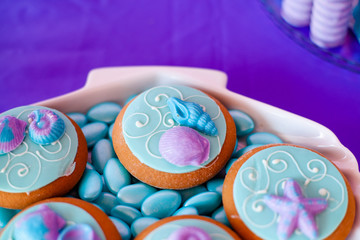 White shell plate full of chocolate candies with blue icing, cookies with shells, stars on the table with shining cloth. Summer season delicious on the party. Sea time theme on the candy bar