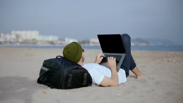 Young millennial generation z man, writes code or responds to emails, uses laptop to work remotely from office while lays on beach,rests back on backpack for travels.Concept social media digital nomad