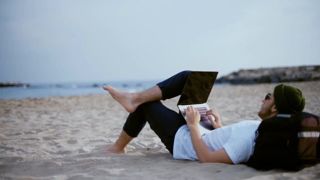 Young millennial remote professional works away from office at beach during vacation or holiday, lays on travel backpack with laptop on lap, types or responds to emails. Copy space for text