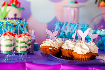 Birthday party concept for girl. Table for kids with cupcakes with withe topind decored purple mermaid tail. Summer season delicious on the party. Sea time theme on the candy bar