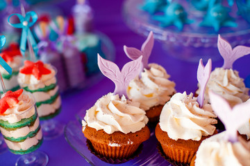 Birthday party concept for girl. Table for kids with cupcakes with withe topind decored purple mermaid tail. Summer season delicious on the party. Sea time theme on the candy bar