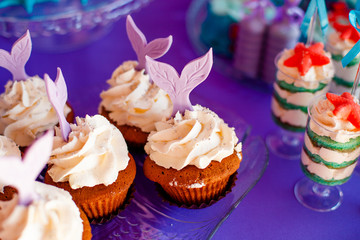 Birthday party concept for girl. Table for kids with cupcakes with withe topind decored purple mermaid tail. Summer season delicious on the party. Sea time theme on the candy bar