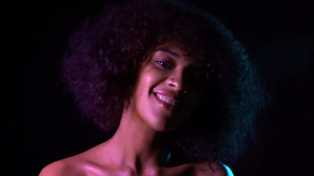 Portrait of young seductive african american girl in pink neon light at black background. Tempting woman with perfect makeup looking at camera and smiling. Glamour, fashion concept.