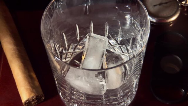 Slow motion of ice cubes being dropped into crystal glass.
Cigar and cutter sit beside glass along with old pocket watch.