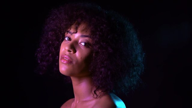 Portrait of young seductive african american girl in pink neon light at black background. Tempting woman with perfect makeup looking at camera and smiling. Glamour, fashion concept.