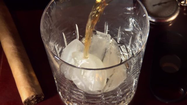 Slow motion of whiskey being poured into glass over ice.
Cigar, cigar cutter and old pocket watch next to glass in shadows.