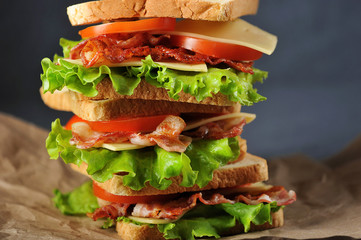 Several sandwiches are laid one on another and occupy the entire space of the frame. Sandwiches with bacon, cheese, slices of tomatoes and green salad leaves. Dark background. Close-up. Macro shooting