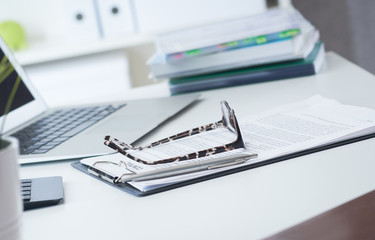 Closeup of white desktop with laptop, spectacles, notepads, pen, smartphone, clipboard.
