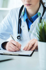 Female doctor filling up prescription form or patient history list at clipboard pad during physical exam or disease prevention while sitting at the desk in hospital closeup.