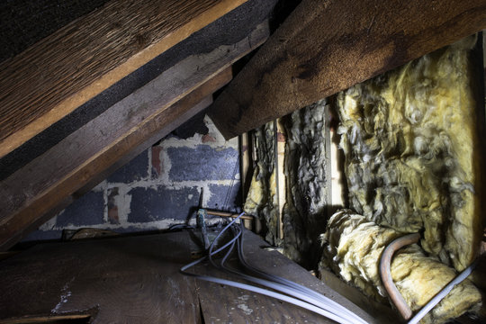 Crawl Space Under The Eves Of A House Showing Old Fibreglass Insulation, Pipework, Rafters, Breezeblock Construction And Old Boarding.