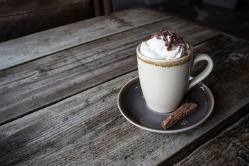 Cup of Hot Chocolate with Squrty Cream and Chocolate Sprinkles on Top.  Flake on saucer.  Standing on rustic wooden table in cafe