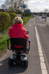 An elderly lady in a red coat enjoying the freedom of an electric mobility scooter alongside a main road