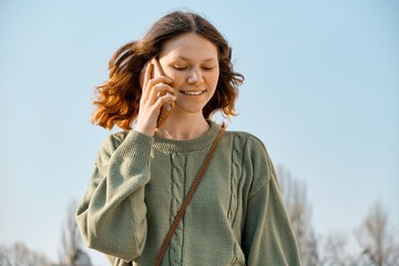 Outdoor portrait of smiling teen girl walking and talking on the phone, blue sky background