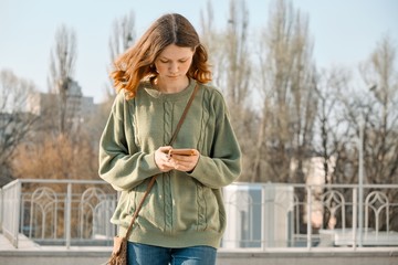 Outdoor portrait of young girl 15 years walking, reading text message on smartphone, spring sunny...