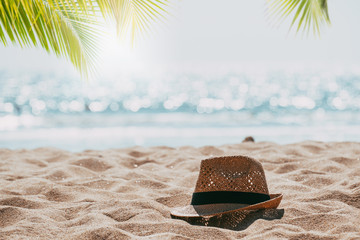 Straw hat on sand tropical beach seascape with palm tree and blur bokeh light of calm sea and sky background. summer vacation background concept.