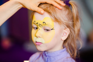 Child animator, artist's hand draws face painting to little girl. Child with funny face painting. Painter makes yellow leopard at girl's face. Children holiday, event, birthday party, entertainment