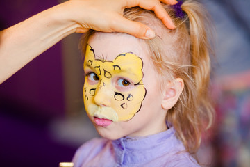 Child animator, artist's hand draws face painting to little girl. Child with funny face painting. Painter makes yellow leopard at girl's face. Children holiday, event, birthday party, entertainment