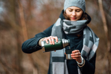 Fototapeta premium Traveler girl pouring tea from thermos, outdoors. Theme travel. Woman pours hot drink in mug from thermos. Drink tea during the hike