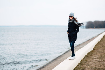 young woman traveler walks along the beach in the fall in windy weather. full length photo of girl in coat, hat, backpack and large scarf goes along the coastline.