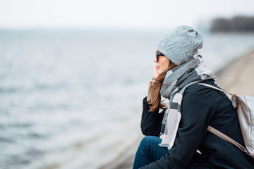 young woman traveler sits near the beach in the fall in windy weather. full length photo of girl in coat, hat, backpack and large scarf outdoors