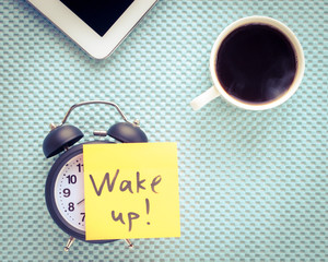 A black alarm clock with a wake up message. Morning coffee and a table to start work. Morning concept.