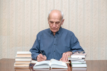 Mature professor works at the table, writes a pen in his notebook, around a lot of books