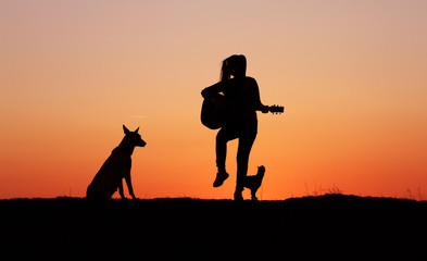 Silhouette girl guitarist on a sunset background, silhouettes of dogs of breed Belgian Shepherd Malinois and miniature pinscher, happy friends, outdoor