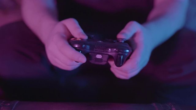 Guy tapping buttons and pushing joystick on a video game controller as he plays in a dark room. Glow of television screen lights scene of man playing video games. Close up of hands.