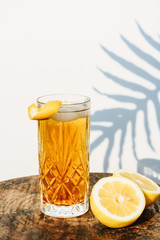 Ice tea in a cocktail glass standing on a wooden table in front of a white wall  outside with shadows of a palm leaf plant in the background